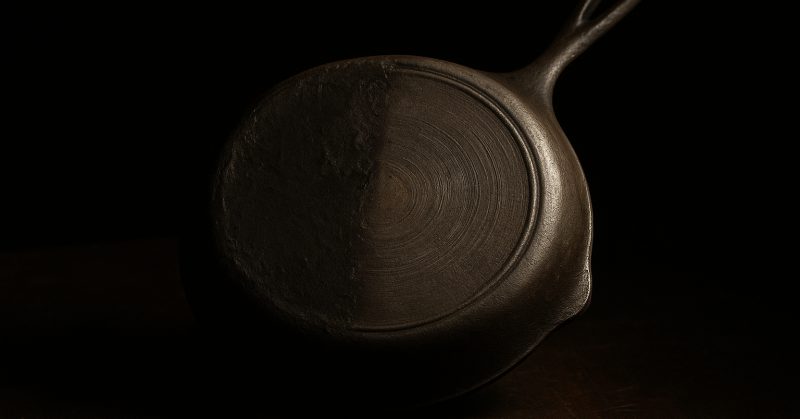 A cast iron skillet part way through stripping, half bare iron and half dark carbon